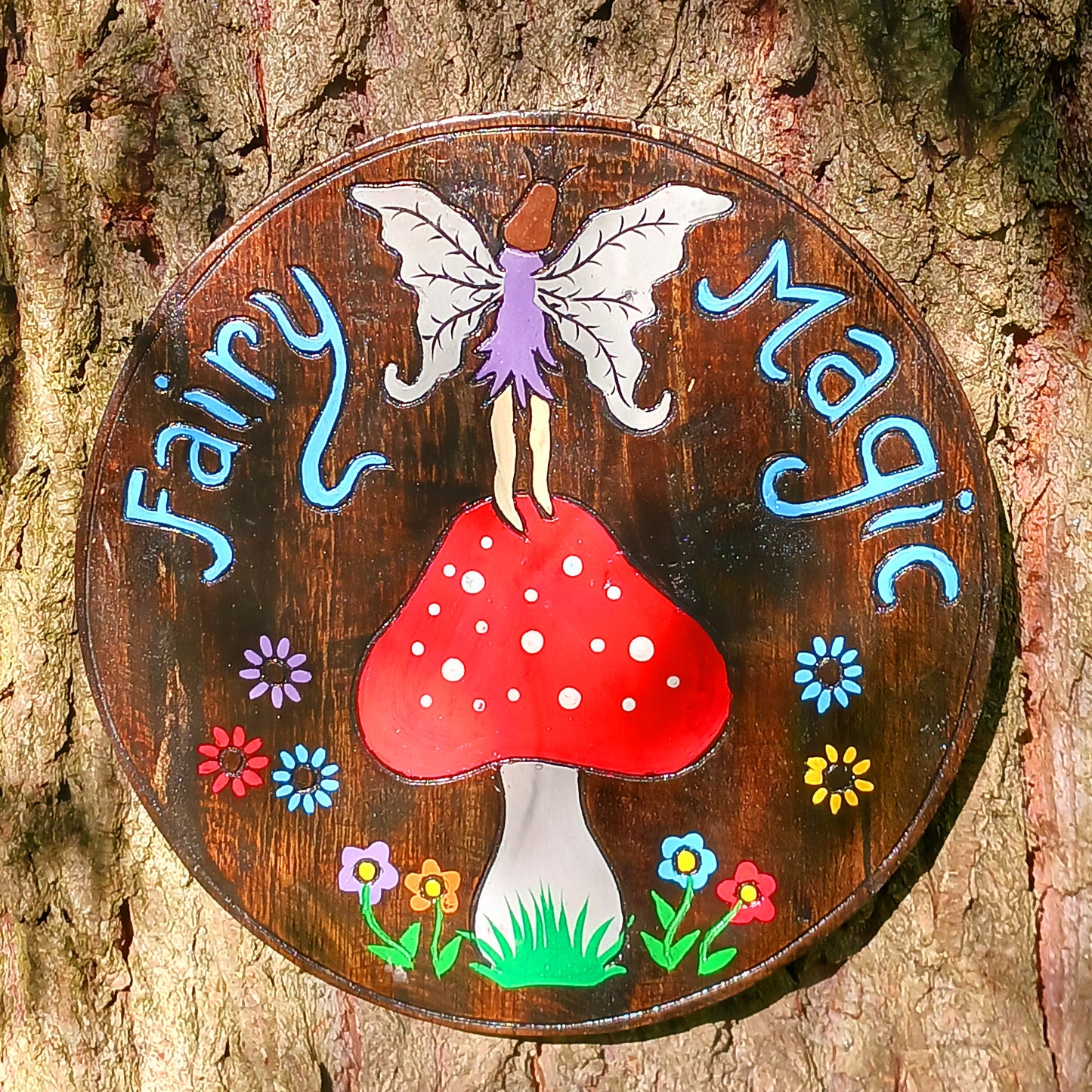 Decorative sign with fairy on a mushroom and flowers, labeled 'Fairy Magic', on a wooden background.
