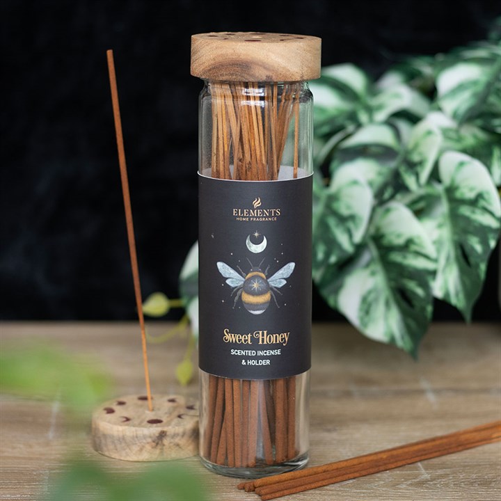 Scented incense sticks in a glass bottle labeled 'Sweet Honey' by Elements on a wooden surface with plants in the background.