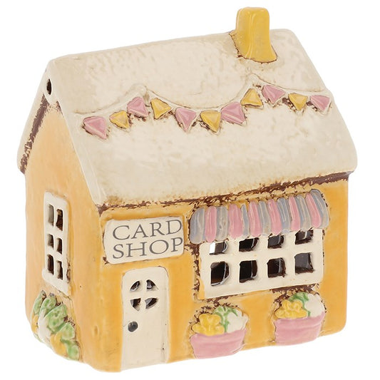 Step through the lantern-lit streets of the village and find the little shop where stories, cards, and gifts are exchanged with a touch of magic. This Village Pottery Cards & Gifts Tealight House is a handcrafted treasure, measuring 14 x 12 x 9cm — the perfect size to bring charm to your hearth or altar. By day, it sits as a quaint ceramic keepsake; by night, a tealight within brings its windows to life, casting a warm and inviting glow. Perfect for witches, dreamers, and lovers of storybook homes, it will 