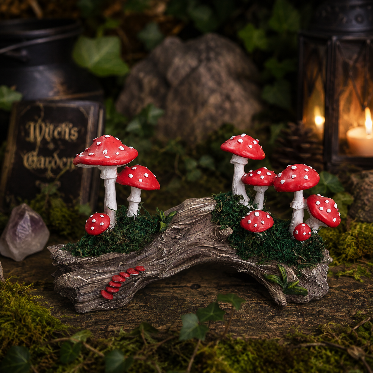 A decorative miniature fairy garden scene featuring a cluster of red and white spotted mushrooms growing on a weathered log with green moss, surrounded by faux foliage, a lantern, and a small sign reading "Witch's Garden.".