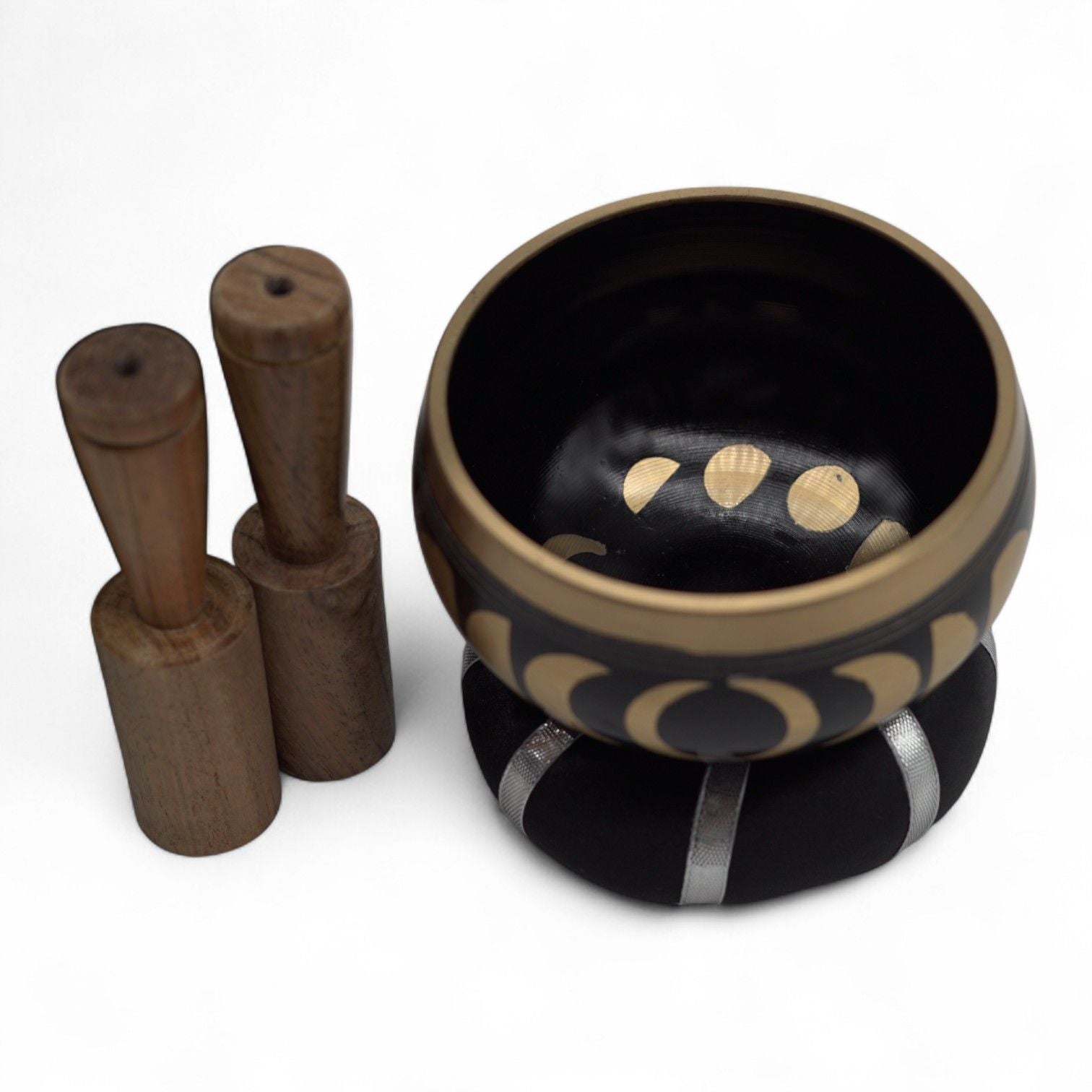A black and gold Tibetan singing bowl set with a wooden mallet and a black cushion, photographed on a clean white background.