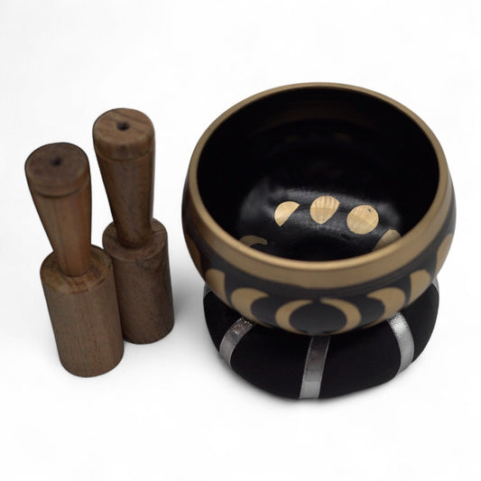 A black and gold Tibetan singing bowl set with a wooden mallet and a black cushion, photographed on a clean white background.