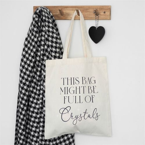 Tote bag with 'This bag might be full of crystals' text hanging on a wooden hook with a scarf and heart decoration.